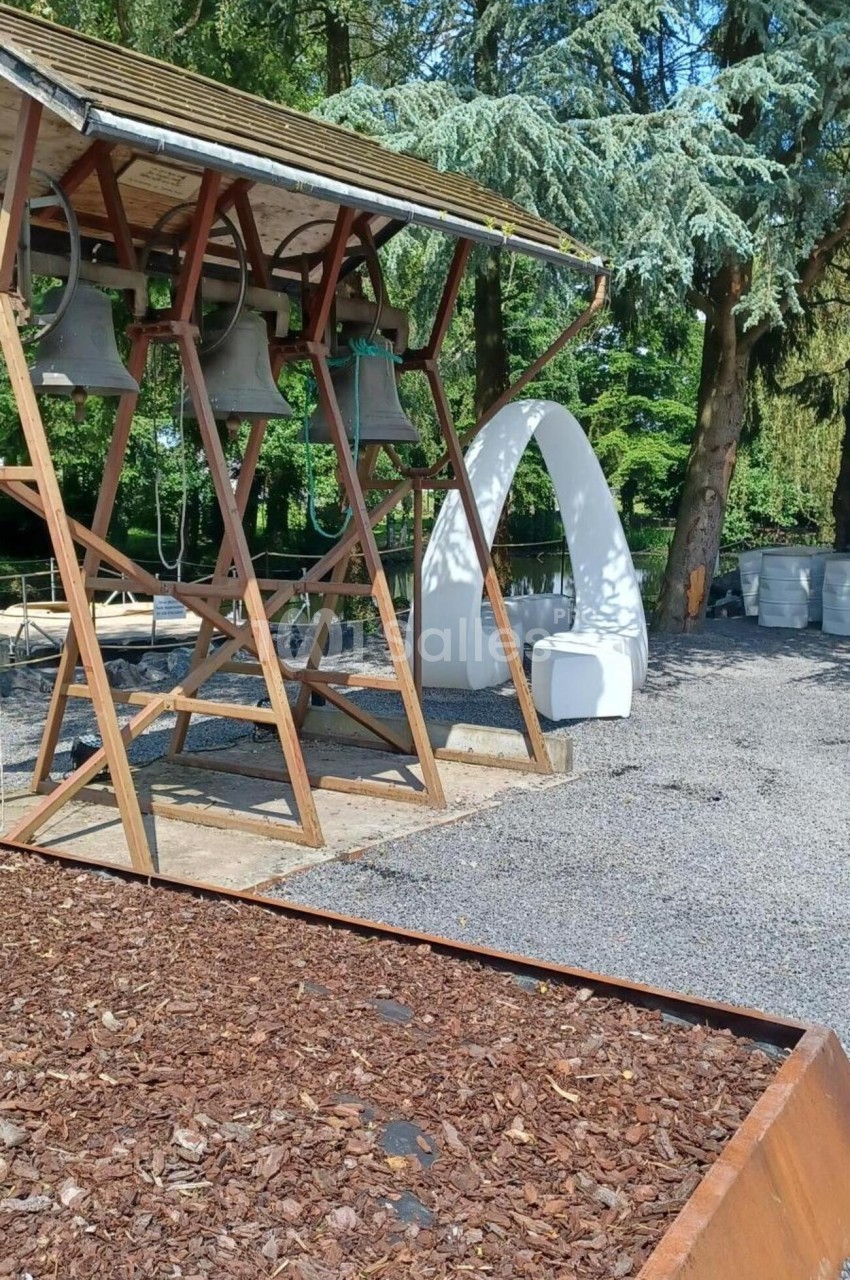 Structure en bois avec des cloches suspendues, entourée d'arbres et d'une arche blanche dans un espace extérieur.