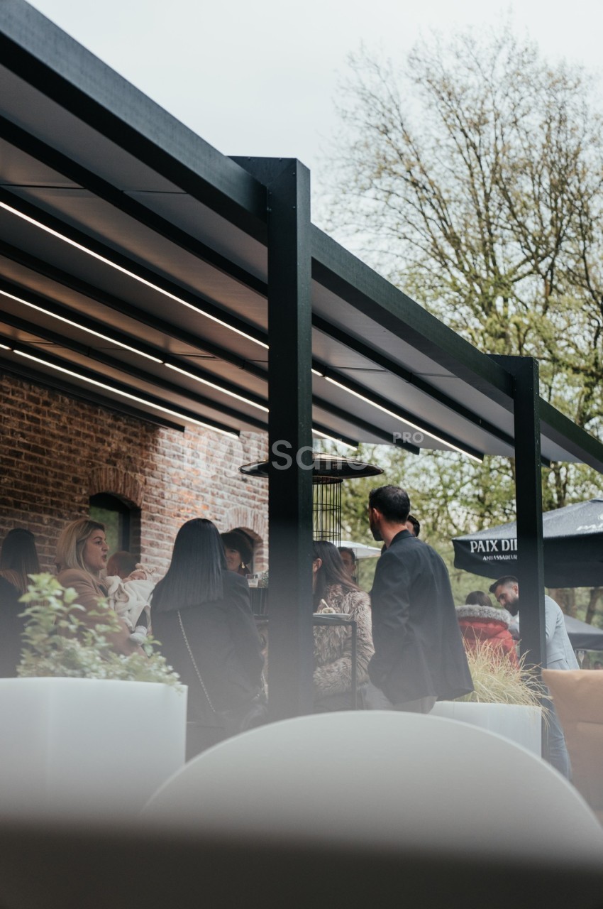 Groupe de personnes discutant sous une pergola moderne près d'un bâtiment en briques, avec des arbres en arrière-plan.