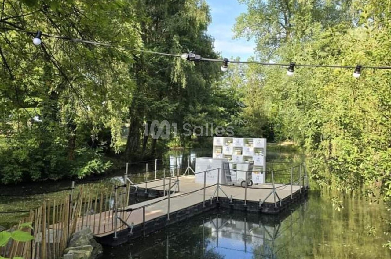 Ponton flottant entouré d'arbres, installé sur un étang calme, avec des guirlandes lumineuses suspendues.