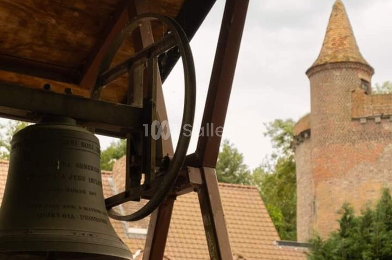 Cloche suspendue sous un toit en bois, avec une tour en briques et des toits en arrière-plan.
