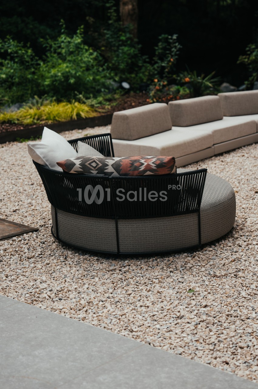 Fauteuil rond en rotin noir avec coussins, placé sur un sol en gravier dans un jardin avec canapé en arrière-plan.