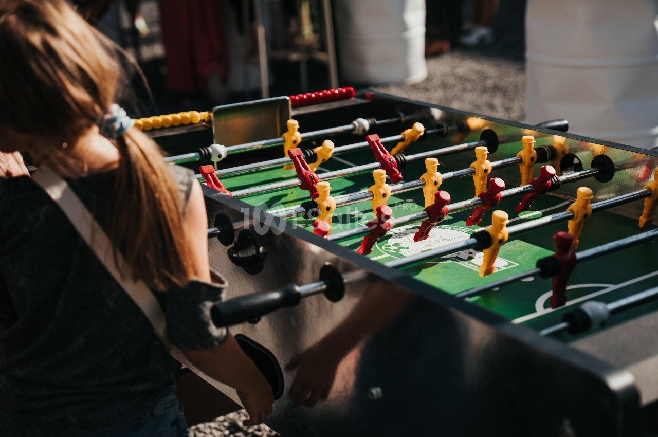 Une enfant joue au baby-foot sur une table en extérieur, entourée d'autres personnes.