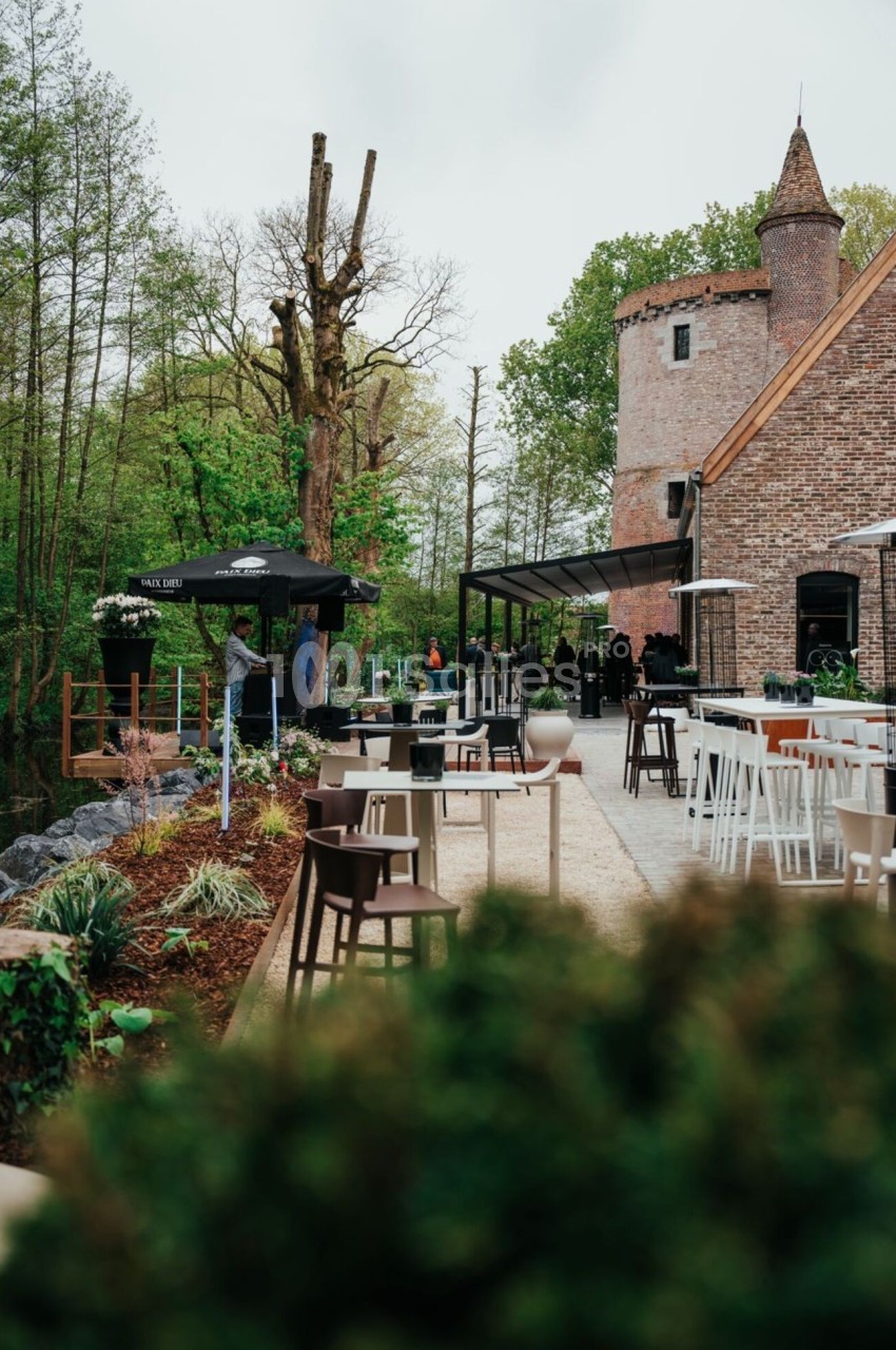 Terrasse extérieure d'un restaurant avec mobilier moderne, entourée de verdure et d'un bâtiment en briques avec une tour.
