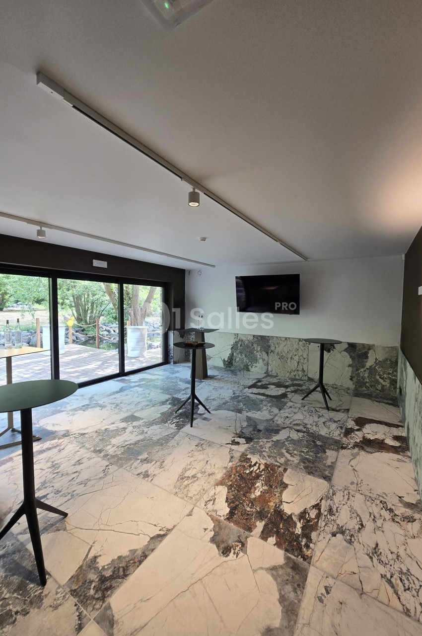 Salle lumineuse avec sol en marbre, tables hautes, écran mural et grandes baies vitrées donnant sur un espace extérieur…