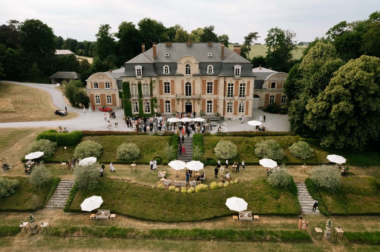 Vue aérienne d'un château entouré de jardins, avec des invités rassemblés devant l'entrée principale.