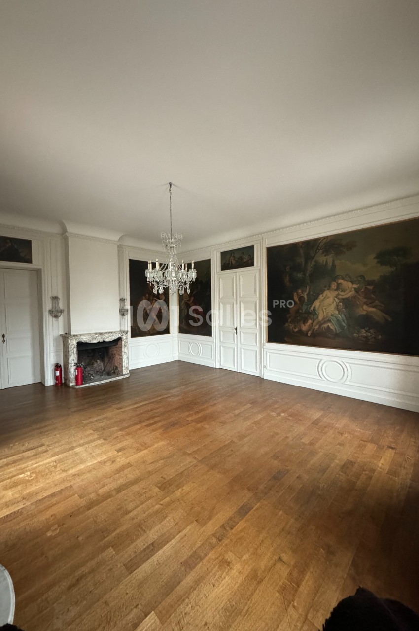 Salle vide avec parquet en bois, murs ornés de grandes peintures, cheminée en marbre et lustre suspendu.