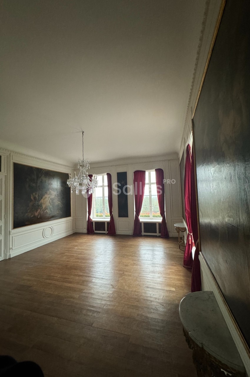 Salle lumineuse avec parquet en bois, murs ornés de tableaux, rideaux rouges et lustre suspendu au plafond.