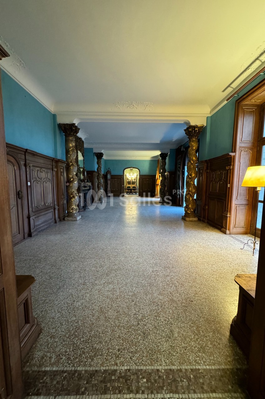 Vue d'un couloir spacieux avec murs bleus, colonnes décoratives, parquet en mosaïque et lumière naturelle entrant par les…