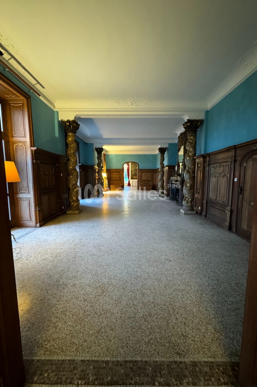 Vue d'un couloir spacieux avec murs bleus, colonnes décoratives, parquet clair et portes en bois sculpté.