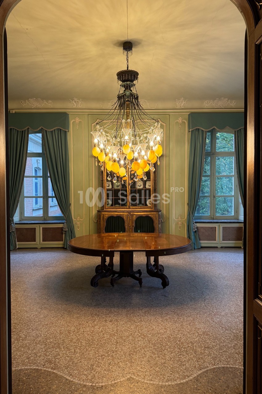 Salle élégante avec un lustre imposant, table ronde en bois, chaises vertes et grandes fenêtres encadrées de rideaux.