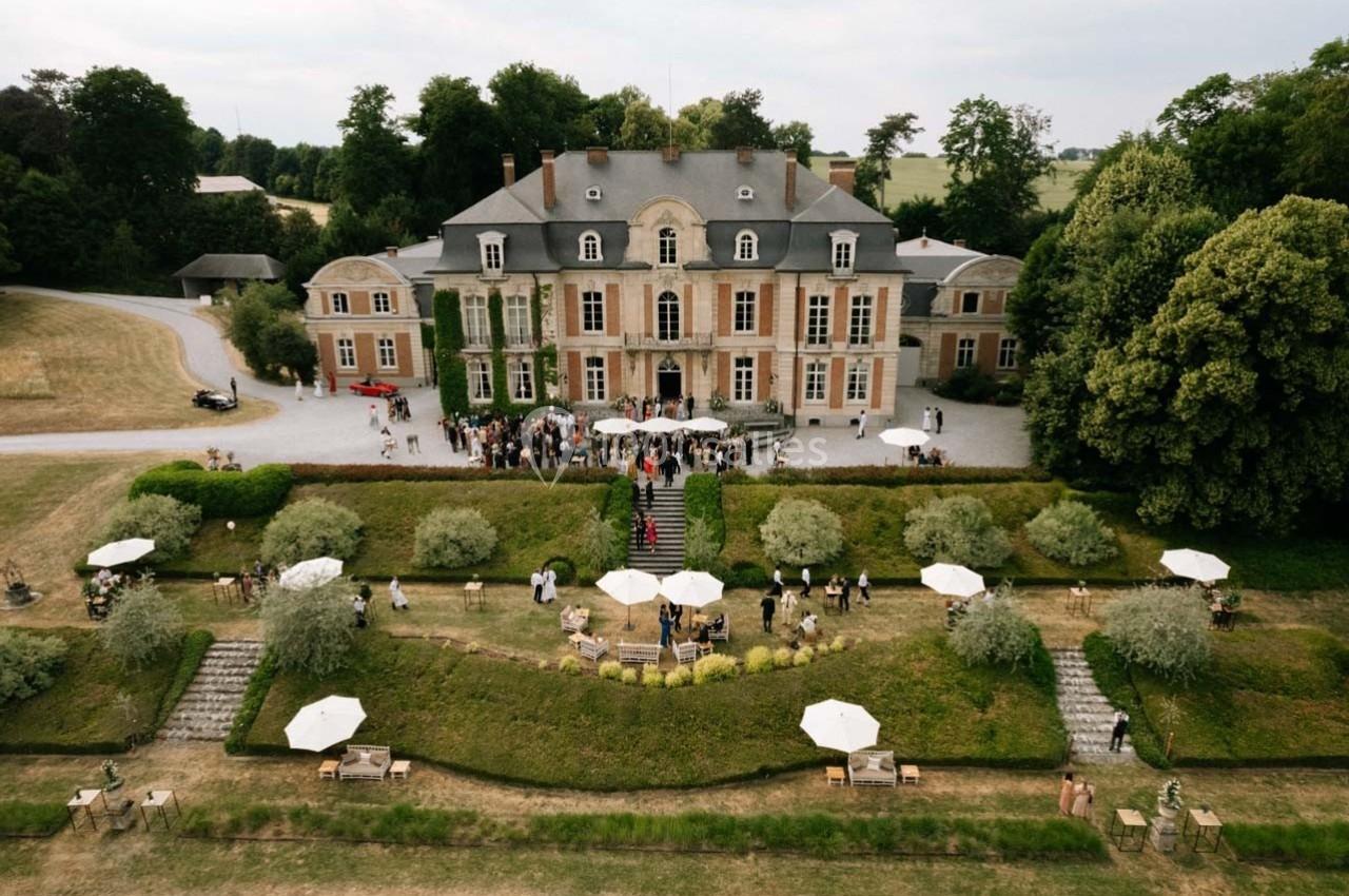 Vue aérienne d'un château entouré de jardins, avec des invités rassemblés pour un événement en plein air.
