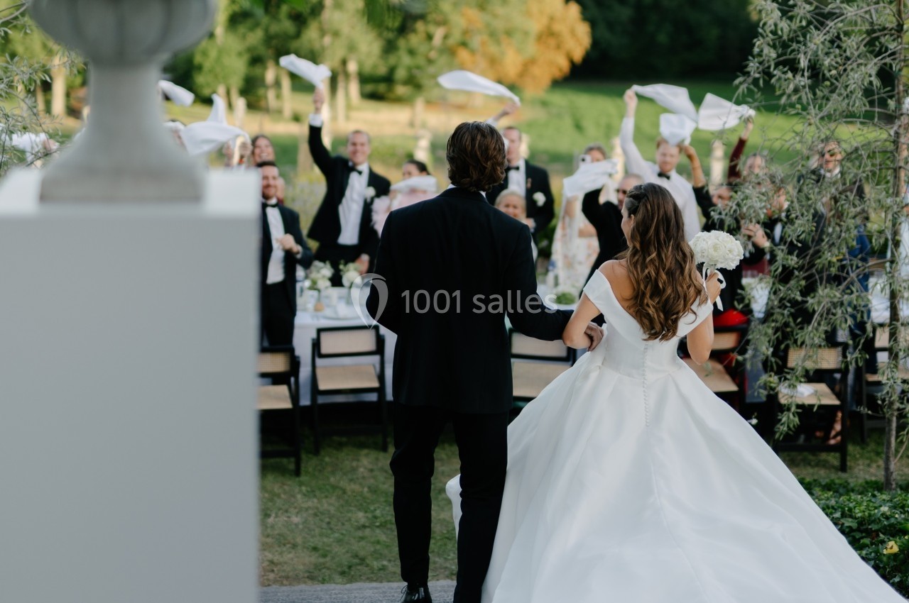 Un couple en tenue de mariage marche vers une table extérieure où des invités agitent des serviettes en l'air.