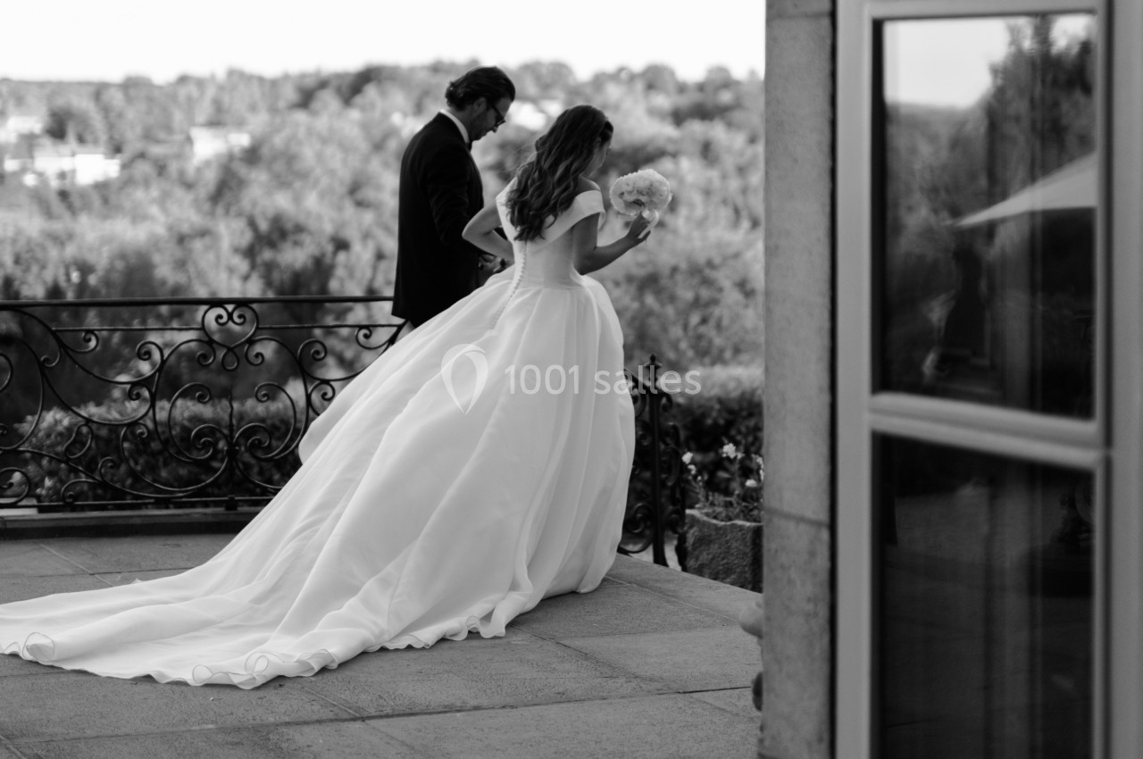 Une mariée en robe longue marche aux côtés d'un homme en costume sur une terrasse avec vue sur un paysage verdoyant.