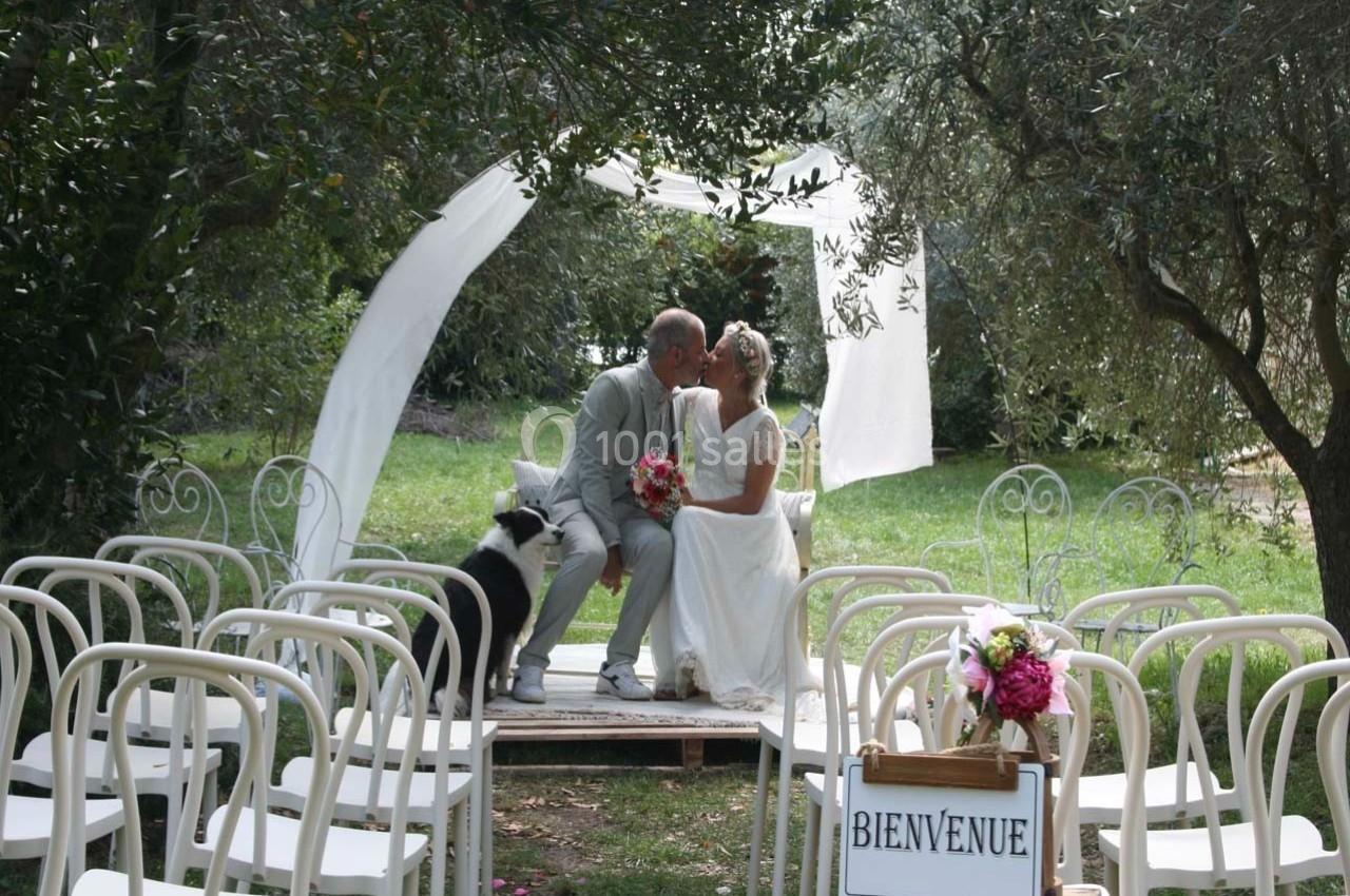 Un couple de mariés s'embrasse sur une petite estrade en plein air, entouré de chaises blanches et d'un chien assis à leurs…