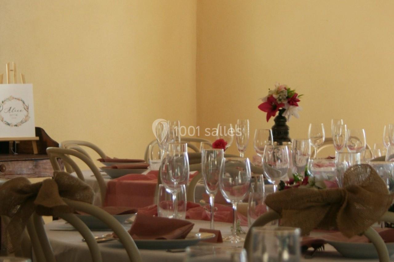Tables dressées avec nappes blanches, serviettes roses, verres à pied et un bouquet de fleurs en centre de table.