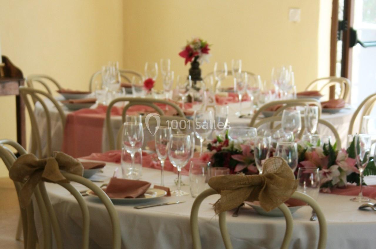 Tables rondes décorées avec nappes blanches, serviettes roses, bouquets de fleurs et verres disposés pour un repas.