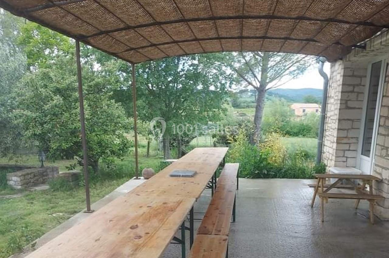 Table en bois avec bancs sous une pergola en osier, donnant sur un jardin verdoyant et un paysage rural.