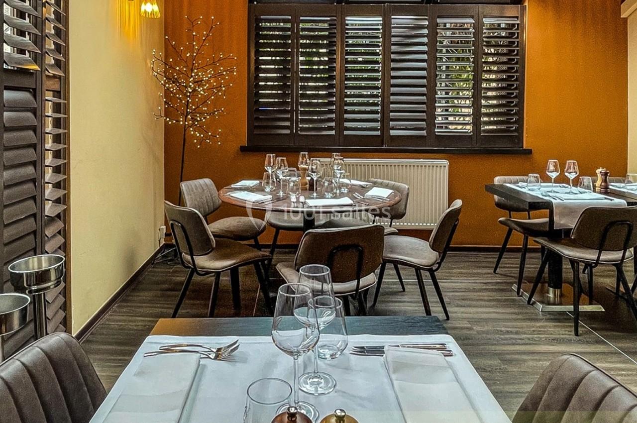 Salle de restaurant chaleureuse avec tables dressées, chaises rembourrées et lumière tamisée près de fenêtres à persiennes.
