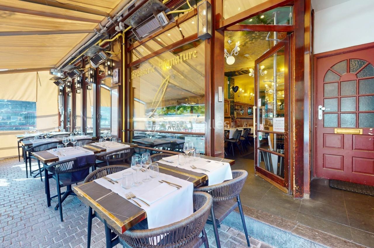 Terrasse d'un restaurant avec tables dressées et napées, donnant sur une entrée vitrée et un intérieur lumineux.