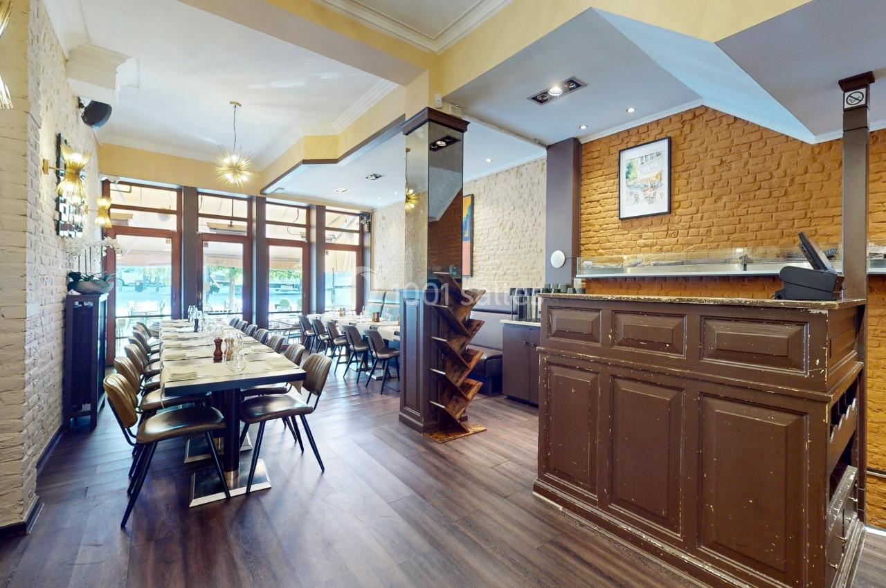 Intérieur d'un restaurant avec tables dressées, chaises alignées, comptoir en bois et murs en briques apparentes.
