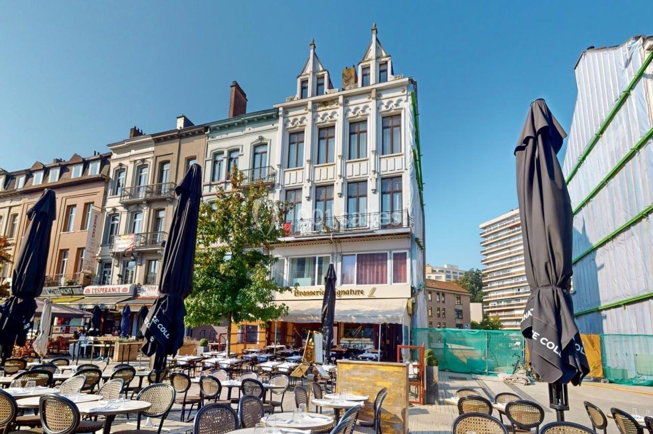 Terrasse de café avec des tables et chaises vides devant des bâtiments anciens et modernes sous un ciel dégagé.