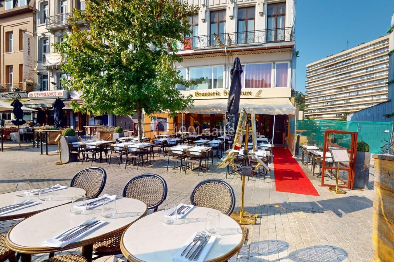 Terrasse d'une brasserie avec tables dressées, située sur une place bordée de bâtiments et d'arbres.