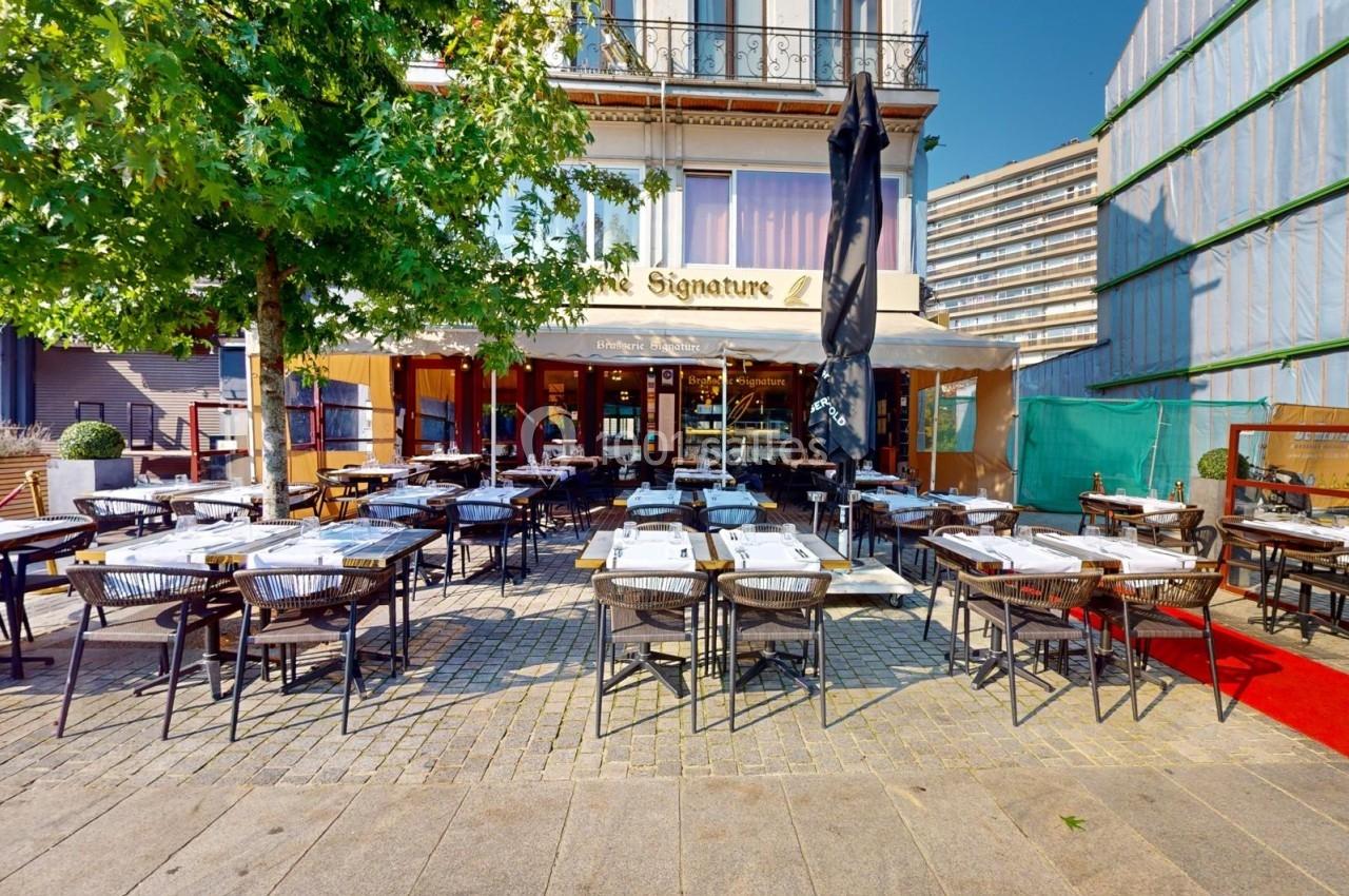 Terrasse d'un restaurant avec des tables dressées, située devant un bâtiment moderne et bordée d'un arbre.