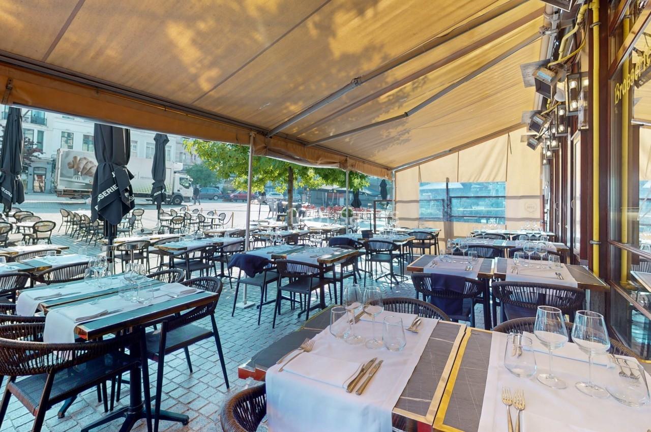 Terrasse de restaurant avec tables dressées sous un auvent beige, donnant sur une place ensoleillée avec des arbres.