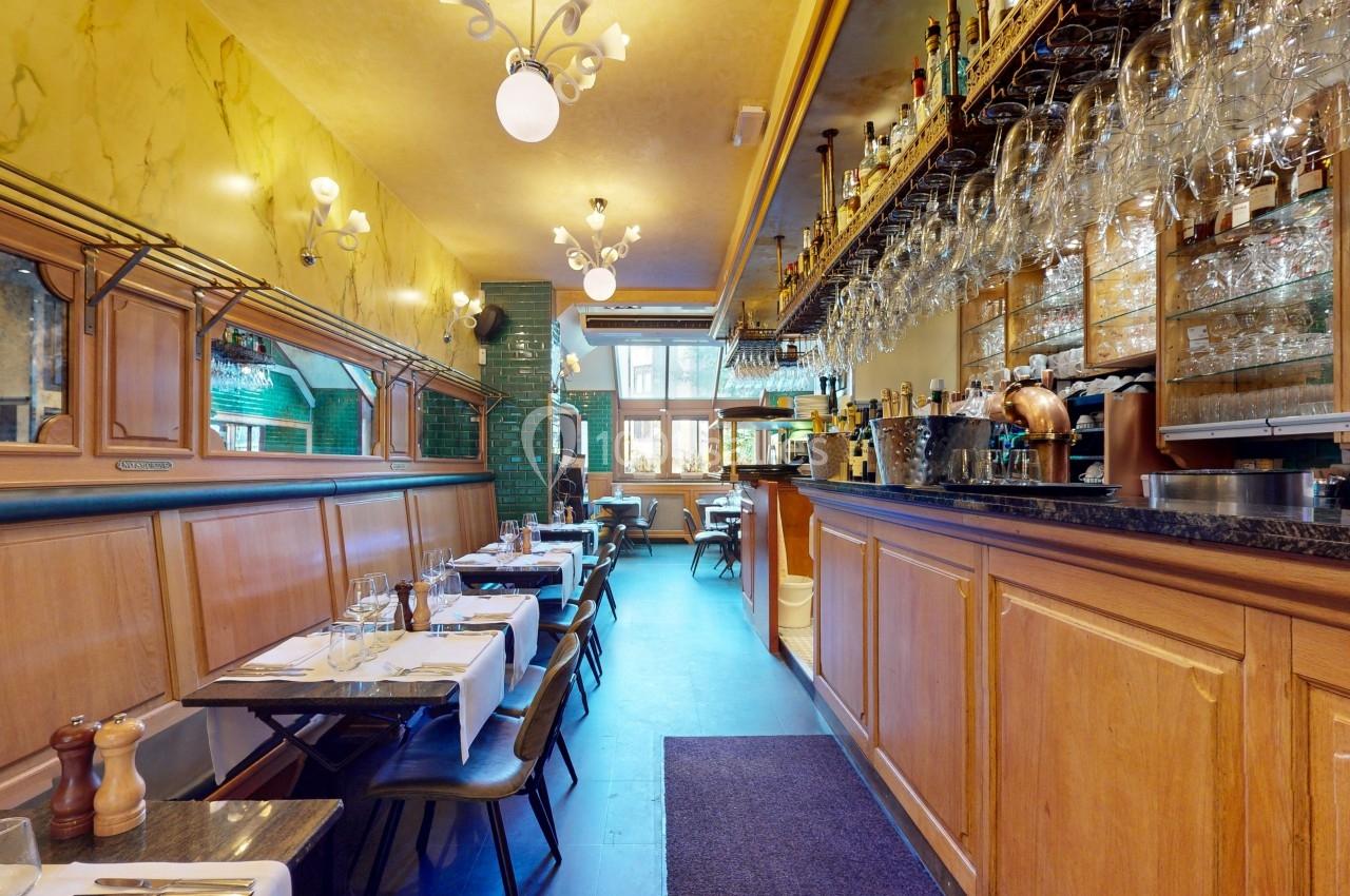 Salle de restaurant avec tables dressées, banquettes en bois, bar en bois et étagères garnies de verres et bouteilles.