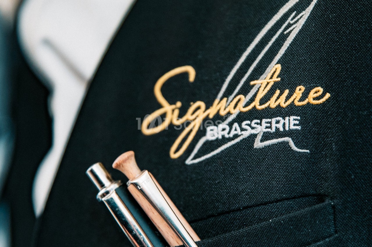 Logo ’Signature Brasserie’ brodé sur un uniforme noir avec des stylos dans une poche.
