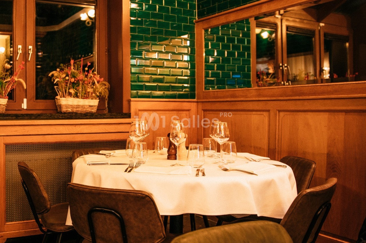 Salle de restaurant avec table ronde dressée, chaises en cuir, murs en bois et carreaux verts brillants.