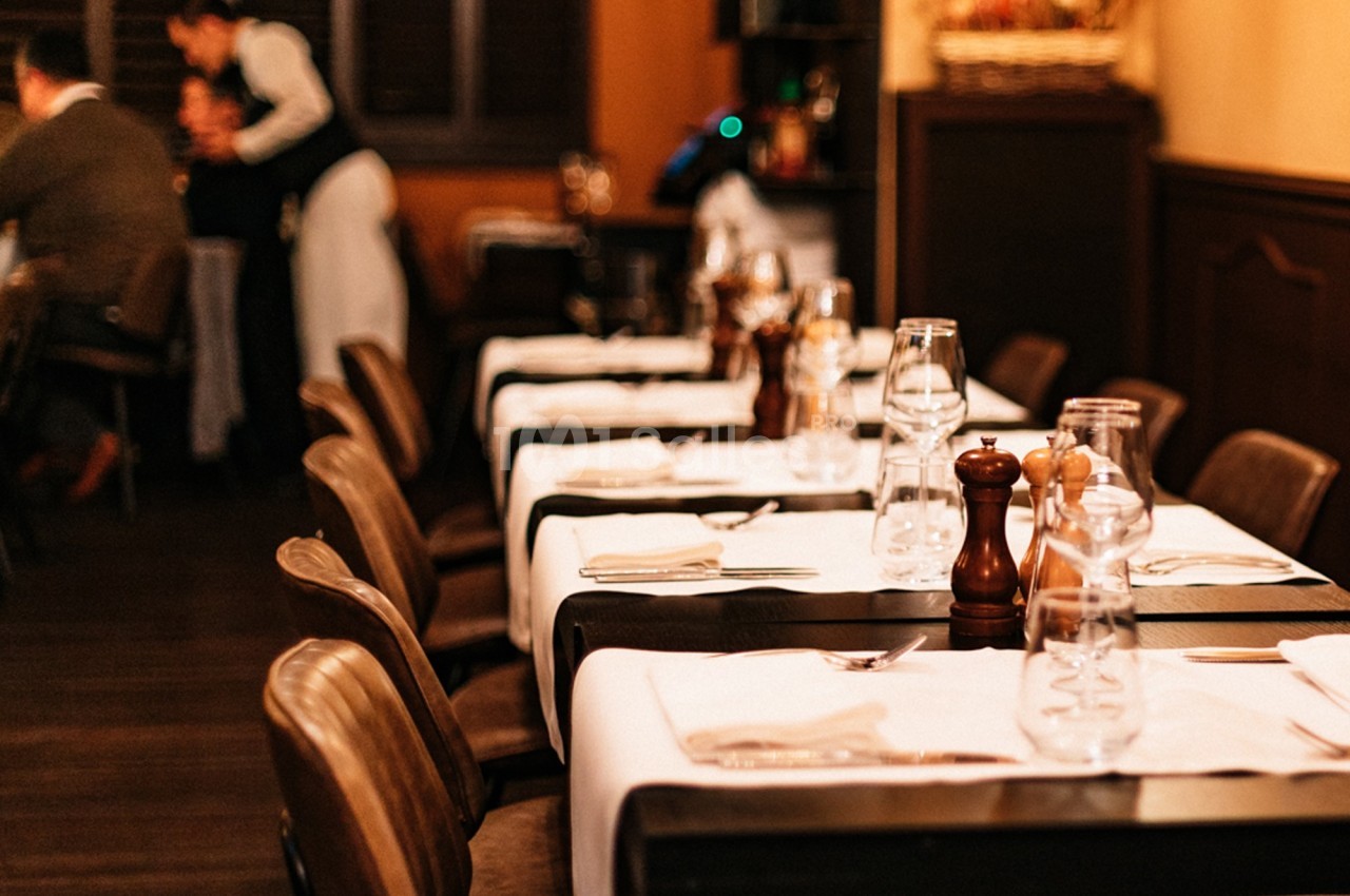 Salle de restaurant avec tables dressées, nappes blanches, verres et condiments, éclairage chaleureux en arrière-plan.