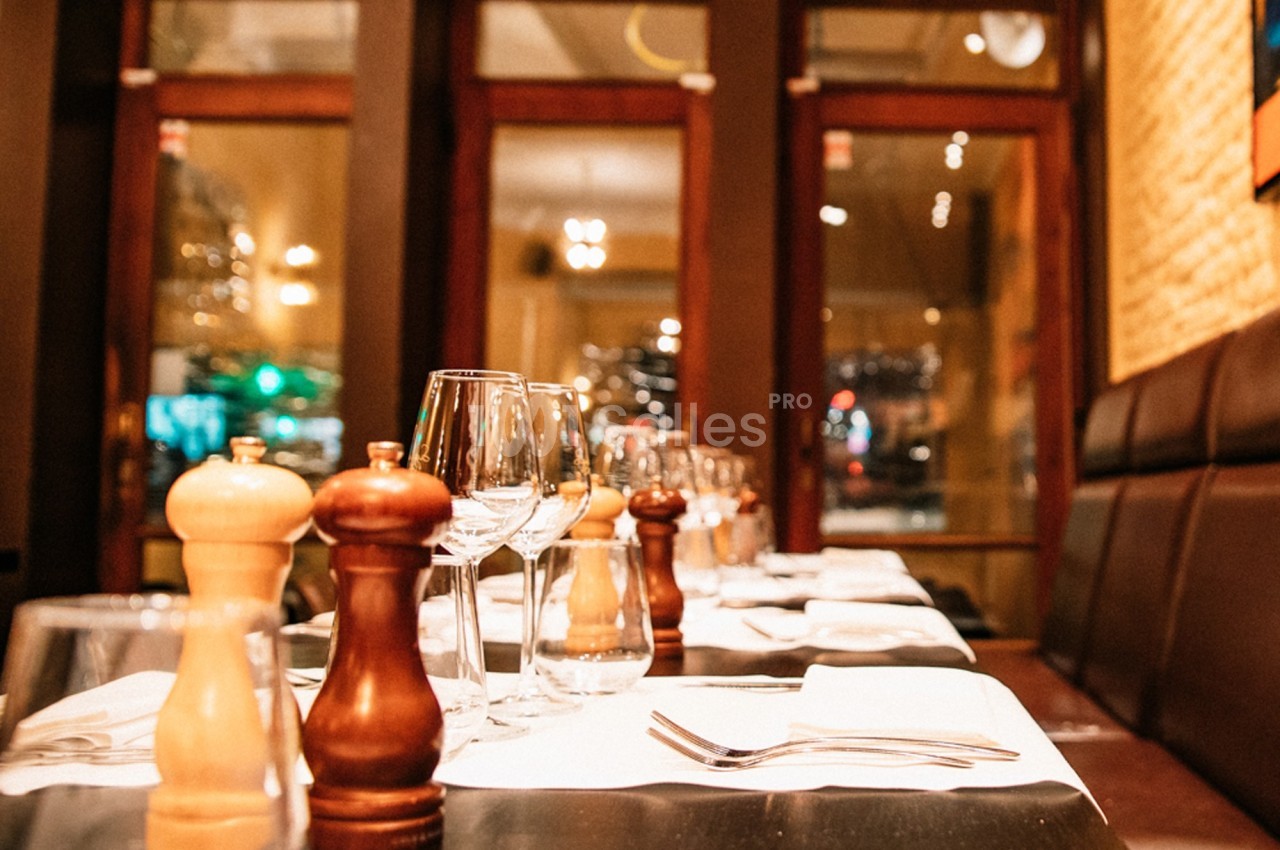 Salle de restaurant avec tables dressées, verres, serviettes et moulins à épices, éclairage chaleureux.