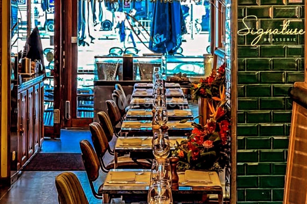 Salle de restaurant avec une longue table dressée, chaises alignées et décoration florale, vue sur l'extérieur en arrière…
