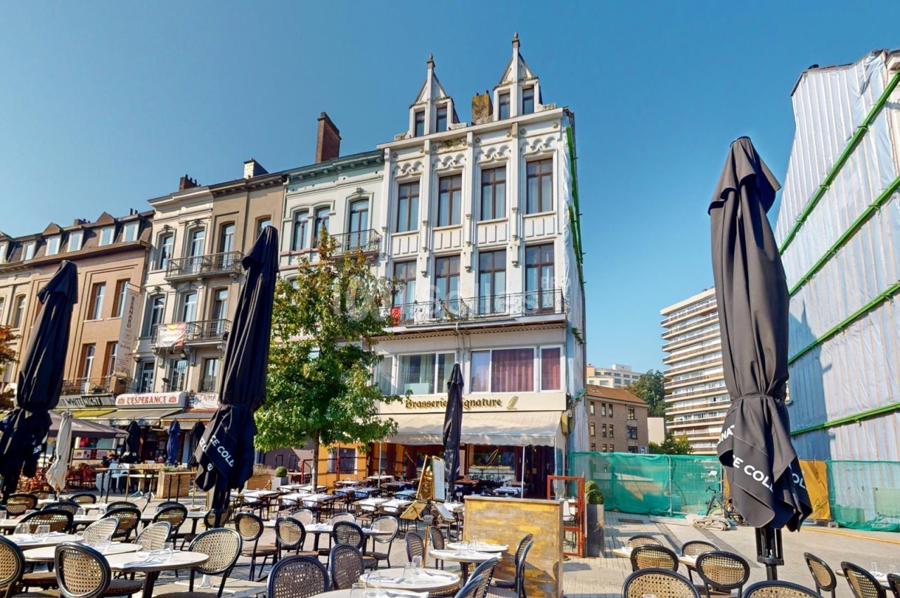 Terrasse de café avec des tables et chaises vides devant des bâtiments anciens et modernes sous un ciel dégagé.