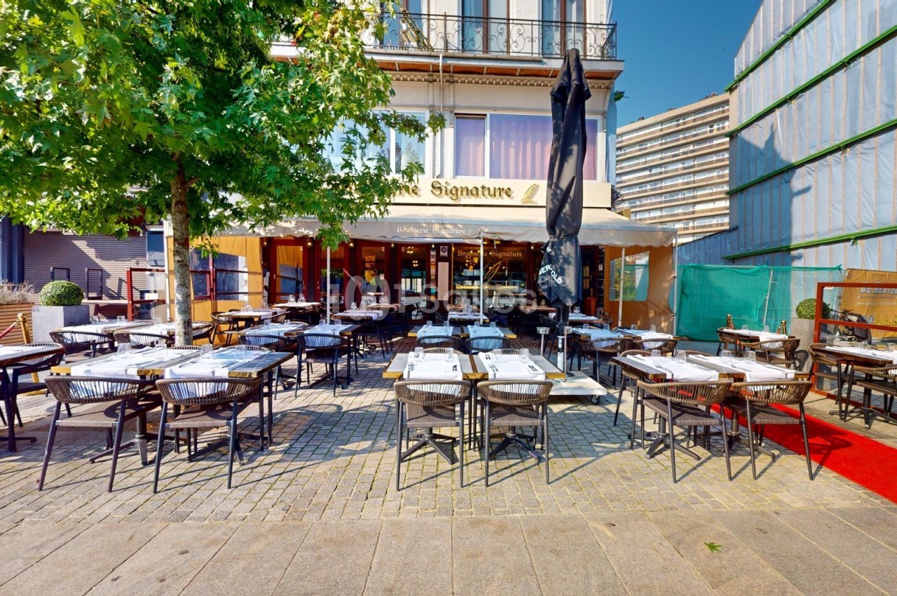 Terrasse d'un restaurant avec des tables dressées, située devant un bâtiment moderne et bordée d'un arbre.