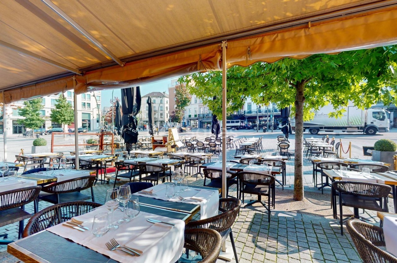 Terrasse de restaurant avec tables dressées sous un auvent, donnant sur une place arborée et des bâtiments en arrière-plan.
