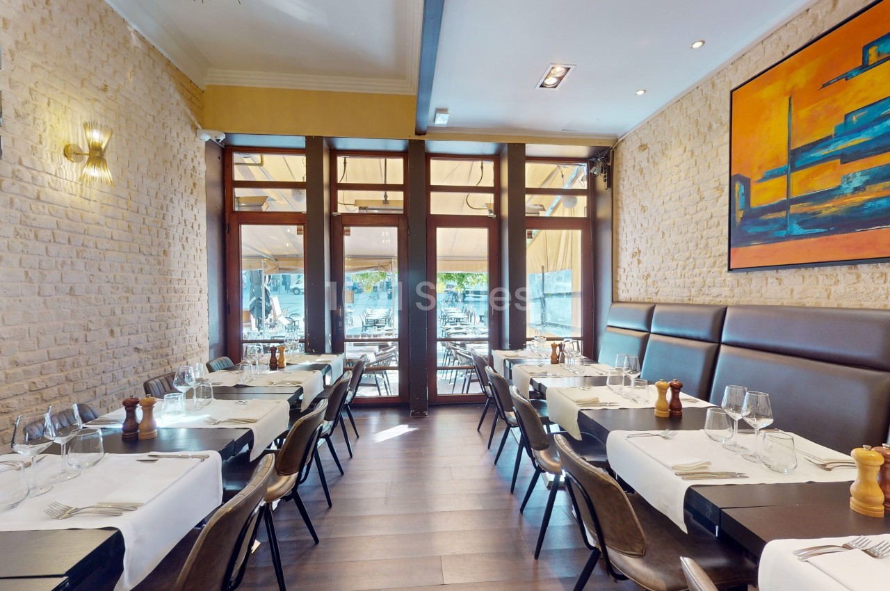 Salle de restaurant lumineuse avec tables dressées, murs en pierre, grandes baies vitrées et tableau coloré au mur.