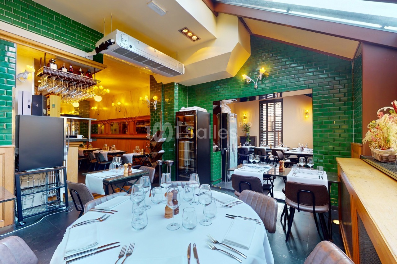 Salle de restaurant lumineuse avec tables dressées, murs en carreaux verts et grandes baies vitrées.