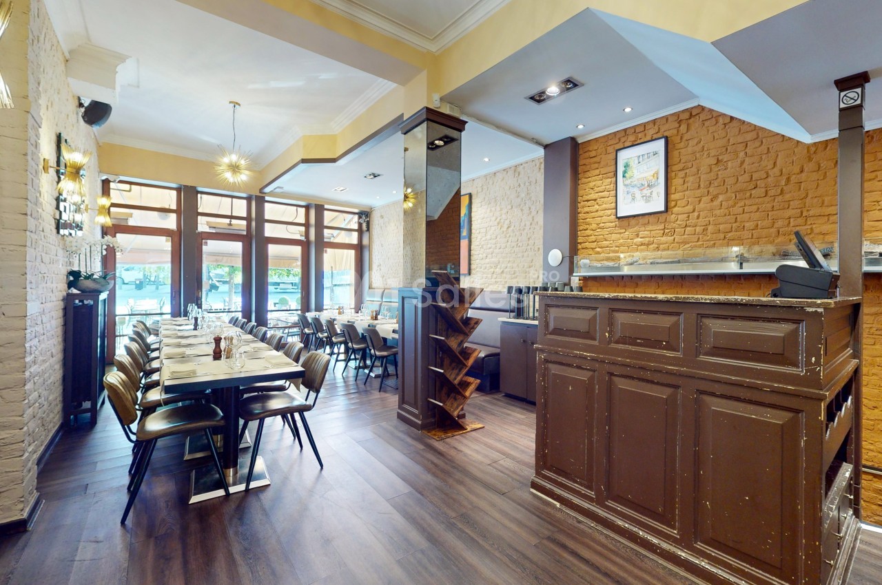 Intérieur d'un restaurant avec tables dressées, chaises alignées, comptoir en bois et murs en briques apparentes.