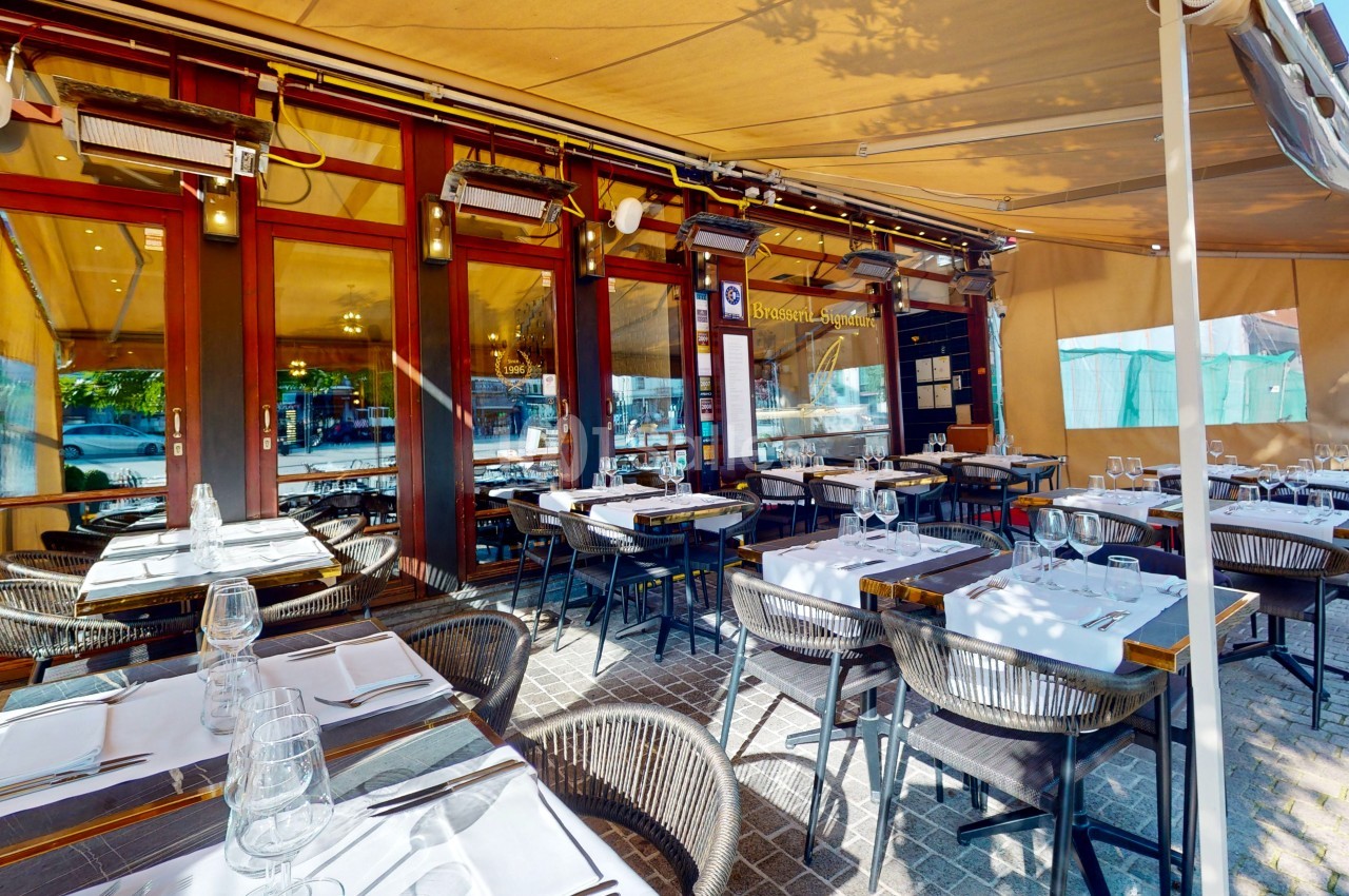 Terrasse d'un restaurant avec tables dressées, chaises en osier et store beige, donnant sur une façade vitrée.