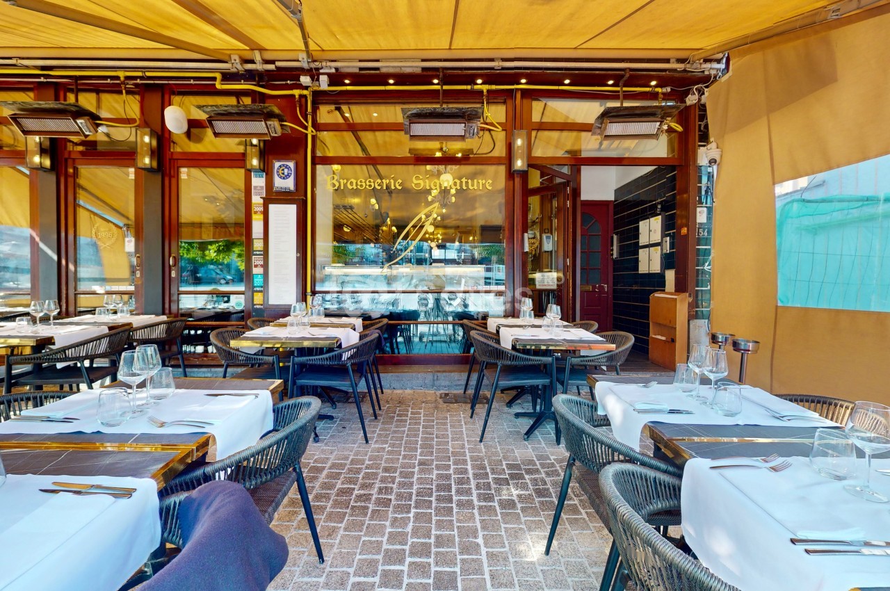 Terrasse d'une brasserie avec tables dressées, nappes blanches et enseigne visible sous un auvent jaune.