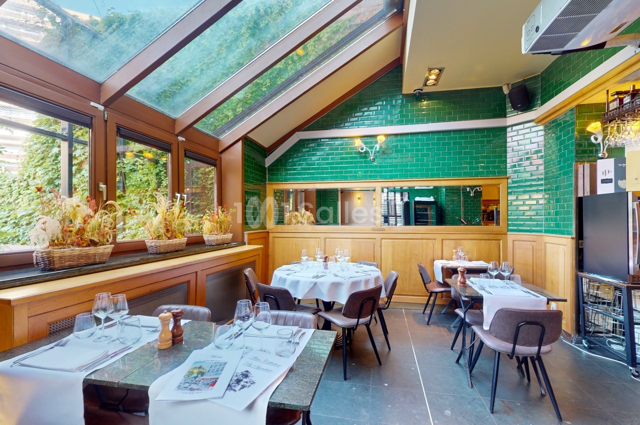 Salle de restaurant lumineuse avec verrière, murs en carreaux verts, tables dressées et plantes en décoration.