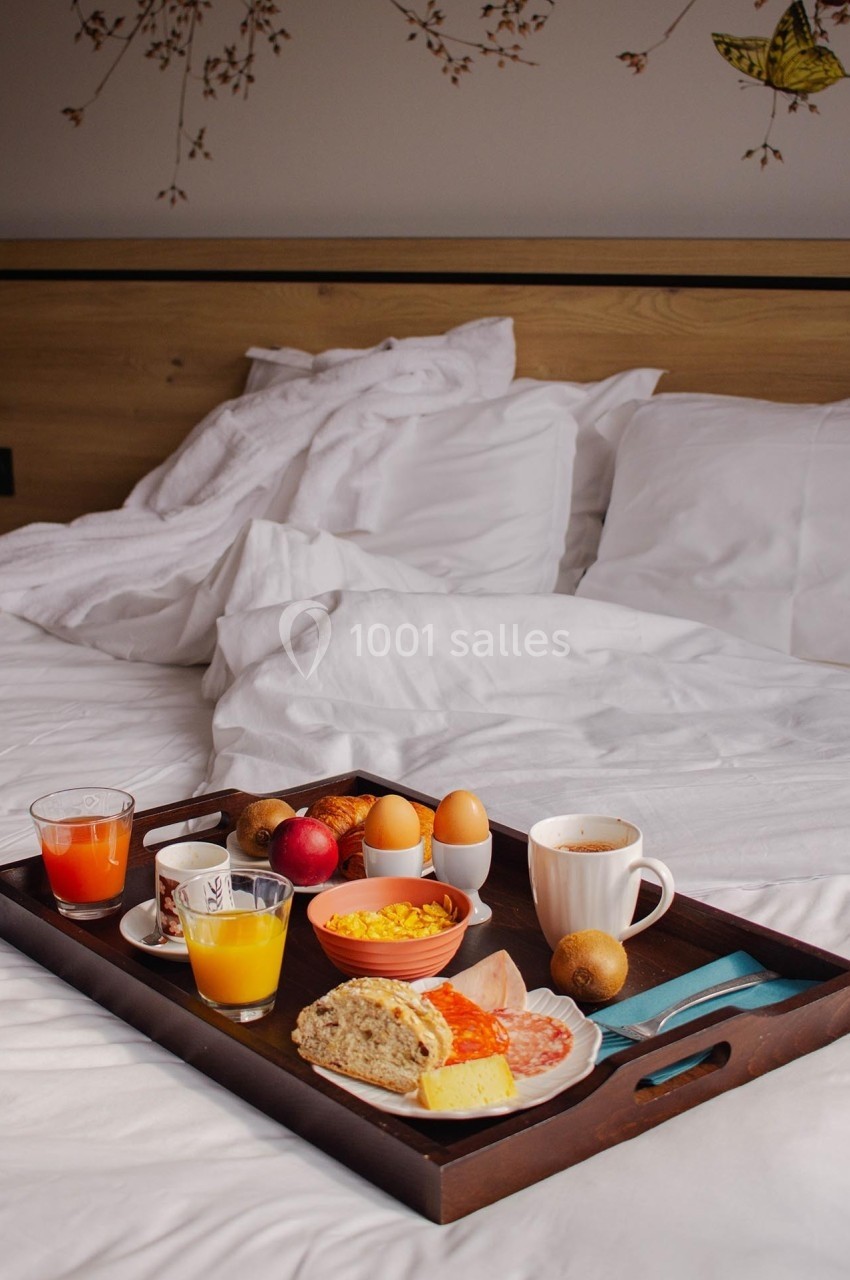 Petit-déjeuner servi sur un plateau avec boissons, viennoiseries, œufs et fruits, posé sur un lit défait.