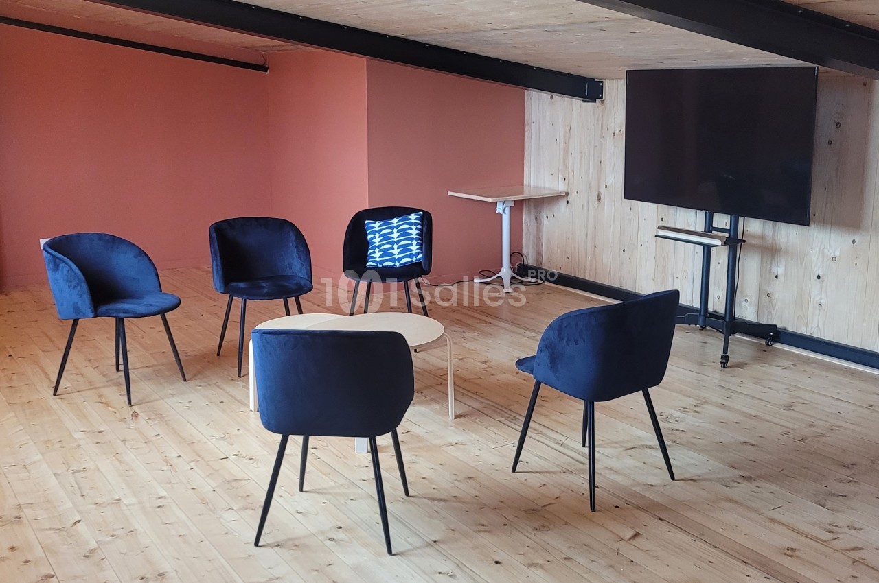 Salle avec parquet en bois, cinq chaises autour d'une table basse, un écran éteint et un extincteur au mur.