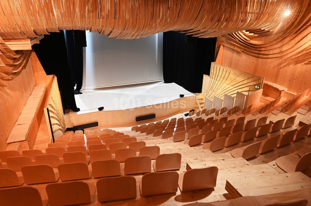 Salle de spectacle en bois avec sièges disposés en gradins et une scène équipée d'un rideau noir.