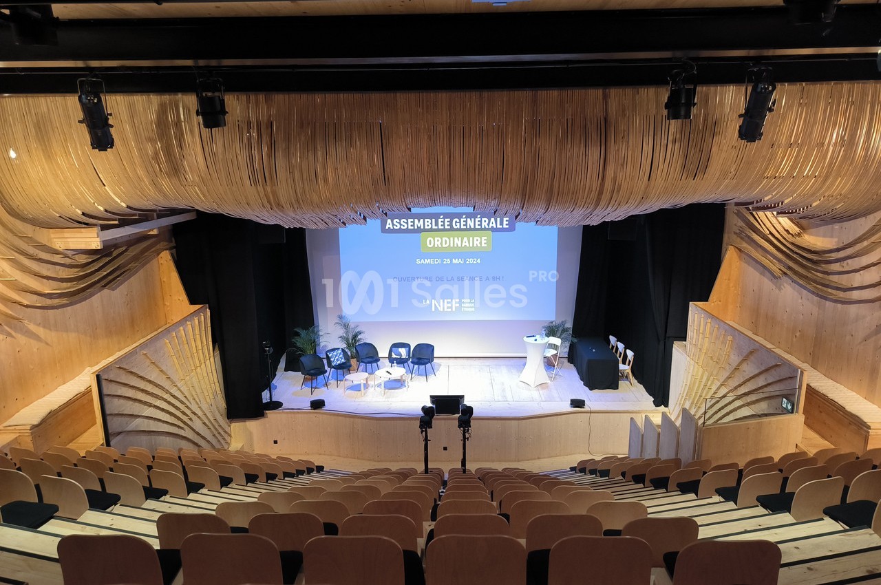 Salle de conférence en bois avec sièges vides, scène équipée de chaises, pupitre et écran affichant ’Assemblée Générale…