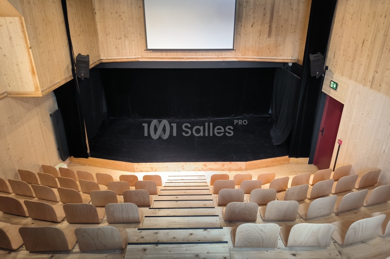 Salle de spectacle en bois avec scène noire, écran blanc suspendu et rangées de sièges en gradins.