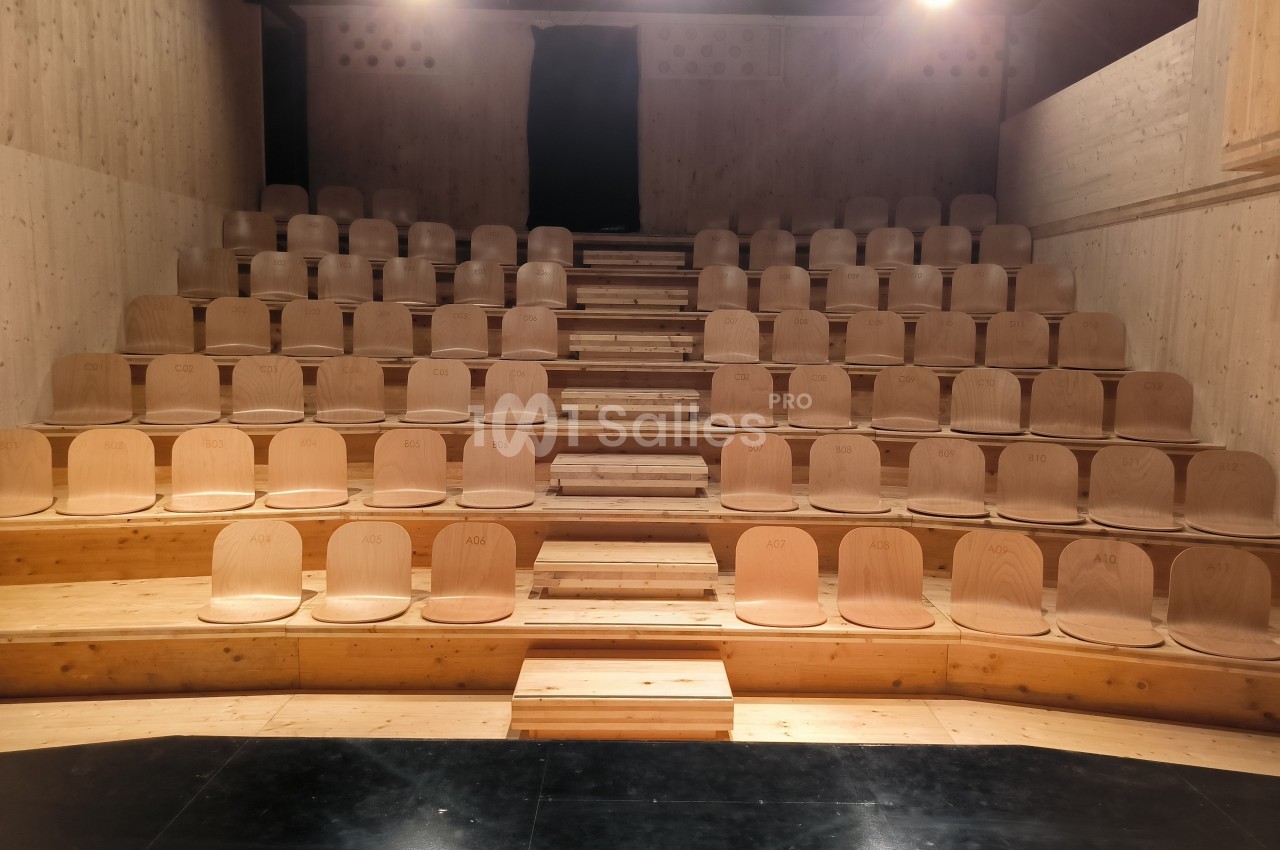 Gradins en bois clair avec des sièges alignés dans une petite salle au sol sombre et éclairée par des projecteurs.
