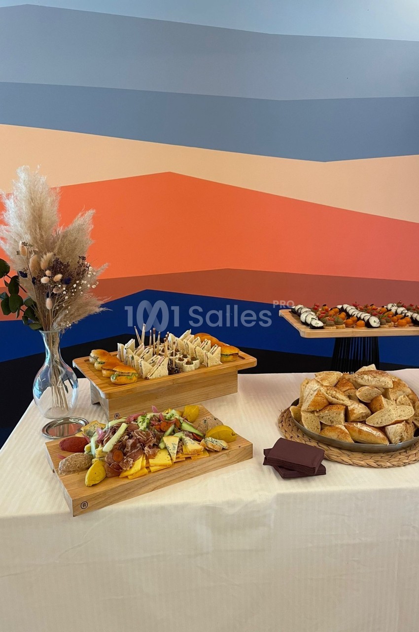 Table dressée avec des plateaux de fromages, charcuteries, sushis et pains, devant un mur aux motifs colorés.